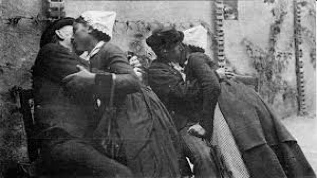 L'image est une photographie en noir et blanc qui semble remonter au XIXe ou au début du XXe siècle. Elle capture un moment intime et quelque peu clandestin entre deux couples. Les couples sont serrés l'un contre l'autre, s'embrassant affectueusement. Les femmes portent des coiffes blanches, typiques d'une époque passée et d'une certaine région géographique. Leurs vêtements sont sombres et semblent être faits de tissus épais, suggérant une certaine pauvreté ou une simplicité vestimentaire. Les hommes sont vêtus de manière similaire, avec des habits sombres et des chapeaux. Les personnages sont de dos ou de profil, et leurs visages ne sont pas parfaitement visibles, ce qui ajoute à la sensation de furtivité et de mystère. L'arrière-plan est flou, mais on distingue un mur de pierre rugueux et une structure architecturale suggérant une ruelle ou un passage étroit. L'éclairage est faible et accentue les contrastes de lumière et d'ombre, contribuant à l'atmosphère quelque peu clandestine et sombre. L'ambiance générale est à la fois romantique et un peu trouble. La proximité physique des couples suggère une passion intense, mais l'obscurité et le manque de détails précis sur les visages laissent planer un sentiment de mystère et d'ambiguïté. Le lieu sombre et isolé renforce l'impression de secret et de transgression éventuelle. L'image suggère un moment volé, un instant d'intimité caché aux regards indiscrets. Le grain de la photographie et le style général suggèrent une image ancienne, qui pourrait être un document historique ou témoigner d'une recherche sur la vie quotidienne à une certaine époque. Texte généré par une IA