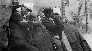 L'image est une photographie en noir et blanc qui semble remonter au XIXe ou au début du XXe siècle. Elle capture un moment intime et quelque peu clandestin entre deux couples. Les couples sont serrés l'un contre l'autre, s'embrassant affectueusement. Les femmes portent des coiffes blanches, typiques d'une époque passée et d'une certaine région géographique. Leurs vêtements sont sombres et semblent être faits de tissus épais, suggérant une certaine pauvreté ou une simplicité vestimentaire. Les hommes sont vêtus de manière similaire, avec des habits sombres et des chapeaux. Les personnages sont de dos ou de profil, et leurs visages ne sont pas parfaitement visibles, ce qui ajoute à la sensation de furtivité et de mystère. L'arrière-plan est flou, mais on distingue un mur de pierre rugueux et une structure architecturale suggérant une ruelle ou un passage étroit. L'éclairage est faible et accentue les contrastes de lumière et d'ombre, contribuant à l'atmosphère quelque peu clandestine et sombre. L'ambiance générale est à la fois romantique et un peu trouble. La proximité physique des couples suggère une passion intense, mais l'obscurité et le manque de détails précis sur les visages laissent planer un sentiment de mystère et d'ambiguïté. Le lieu sombre et isolé renforce l'impression de secret et de transgression éventuelle. L'image suggère un moment volé, un instant d'intimité caché aux regards indiscrets. Le grain de la photographie et le style général suggèrent une image ancienne, qui pourrait être un document historique ou témoigner d'une recherche sur la vie quotidienne à une certaine époque. Texte généré par une IA